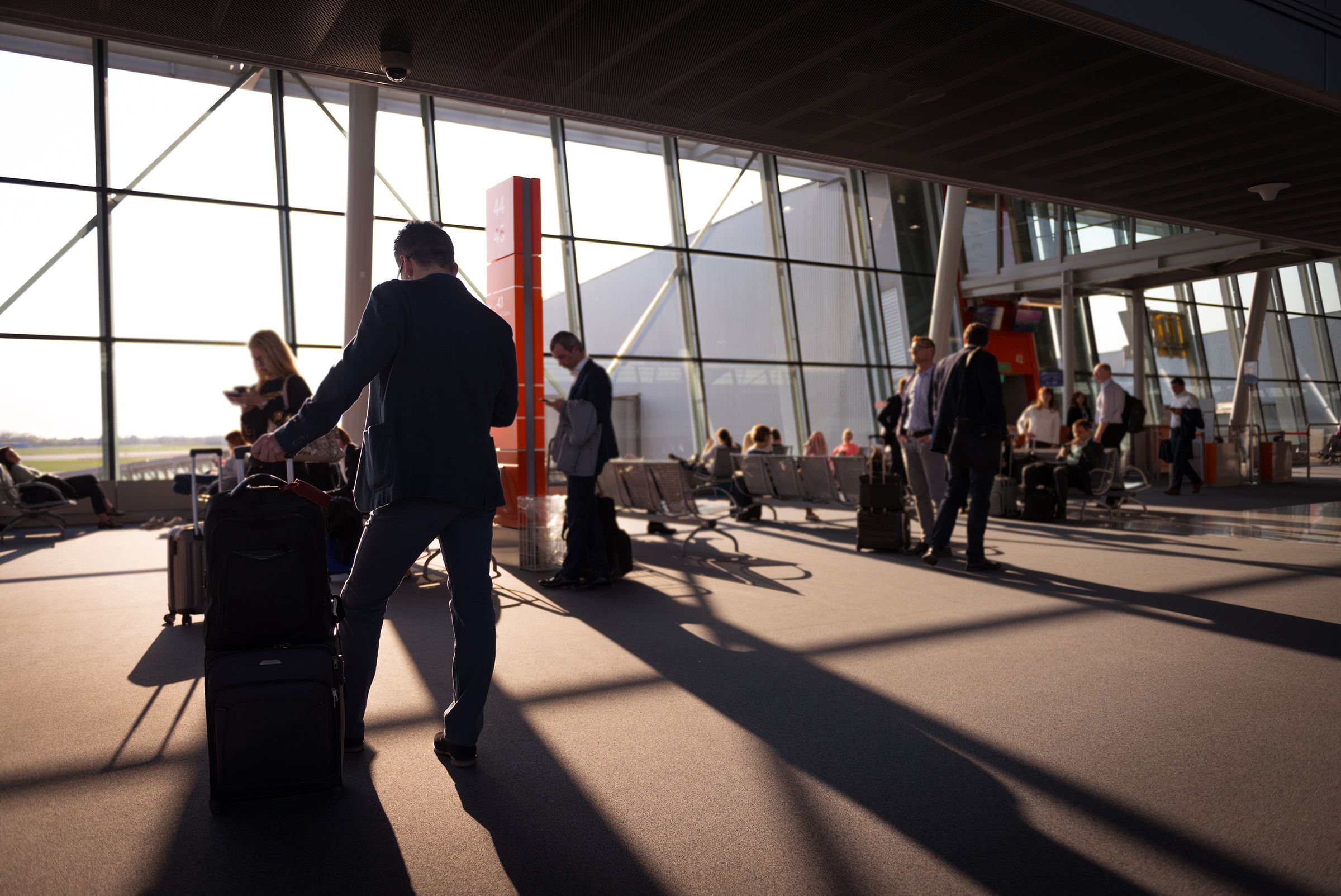 Travelers with rolling luggage inside a sunlit airport terminal.
