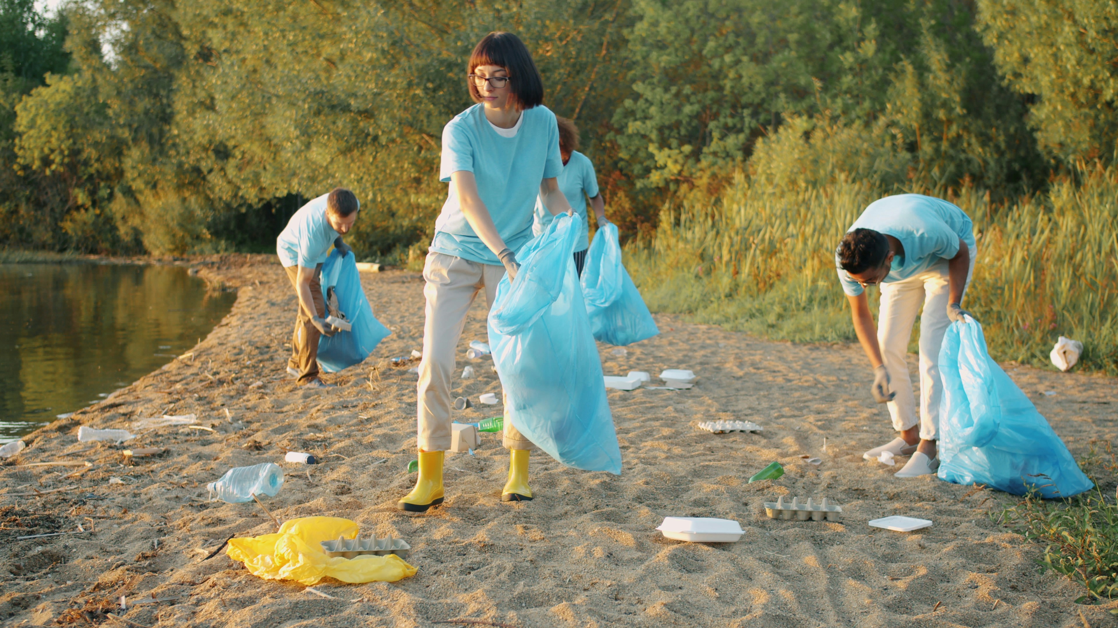 Four volunteers wearing gloves collect litter into blue bags on a sandy shore.