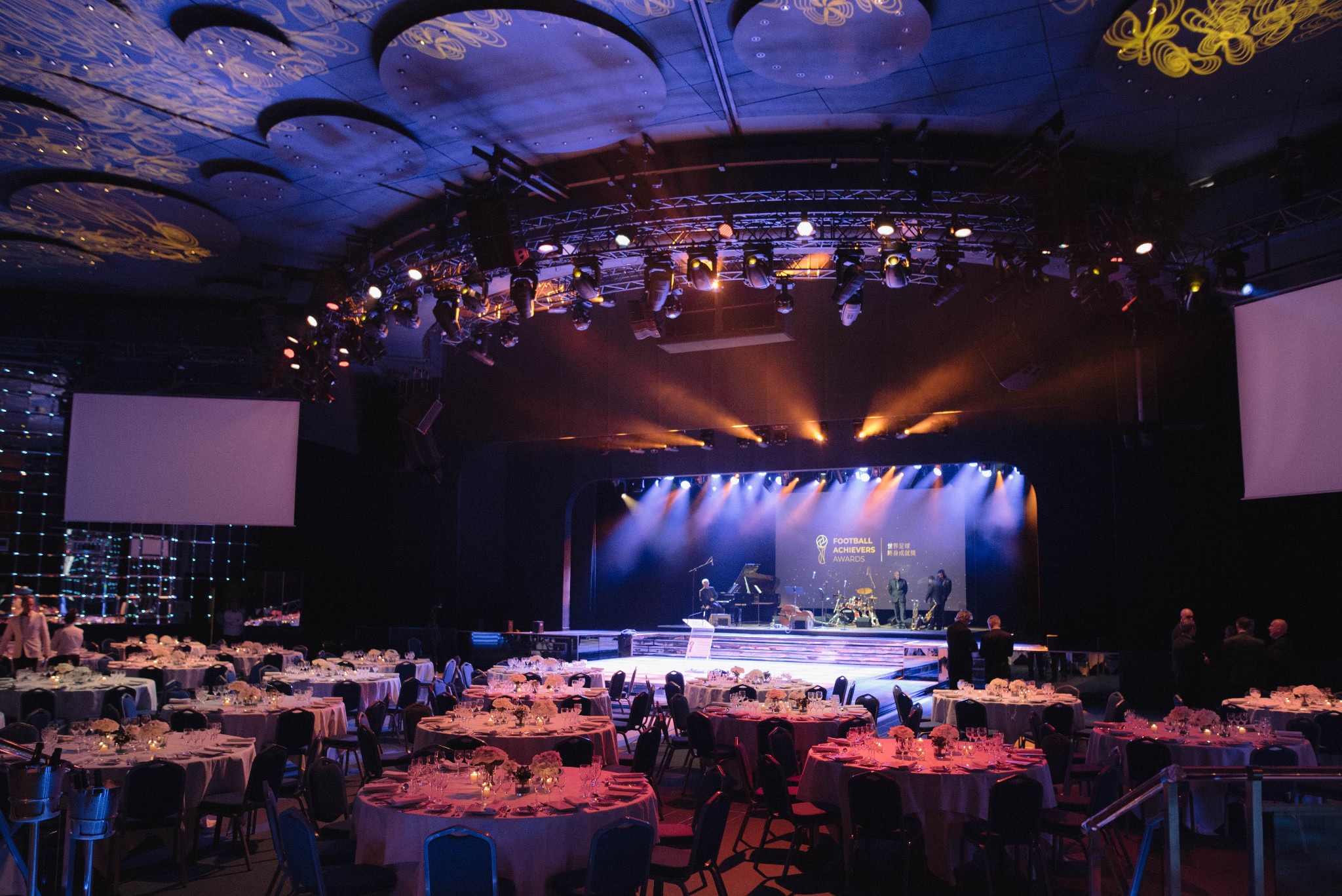 A formal event room with round dinner tables facing an illuminated stage beneath production lighting.