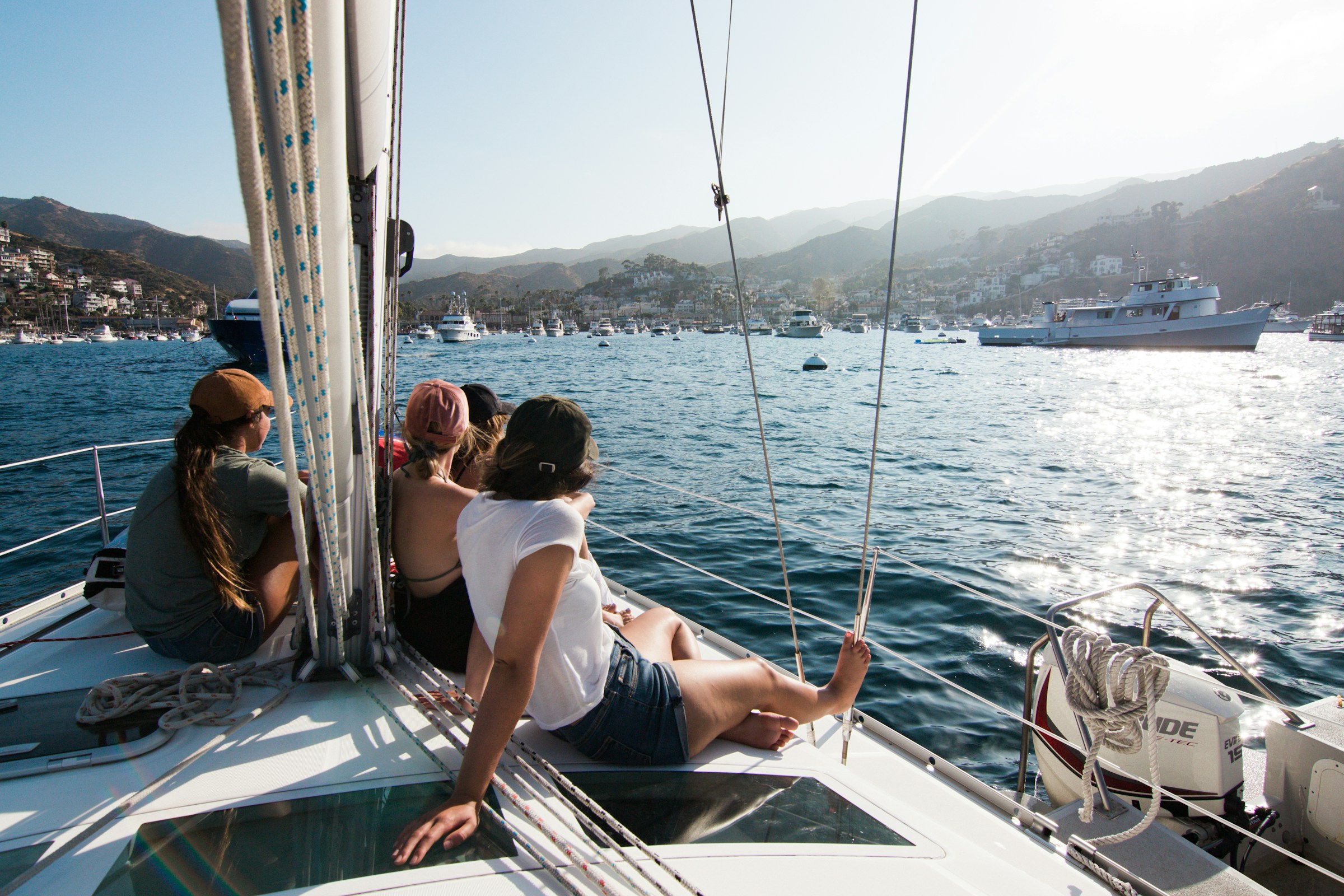 Passengers seated on a sailboat’s foredeck looking across a coastal harbor.