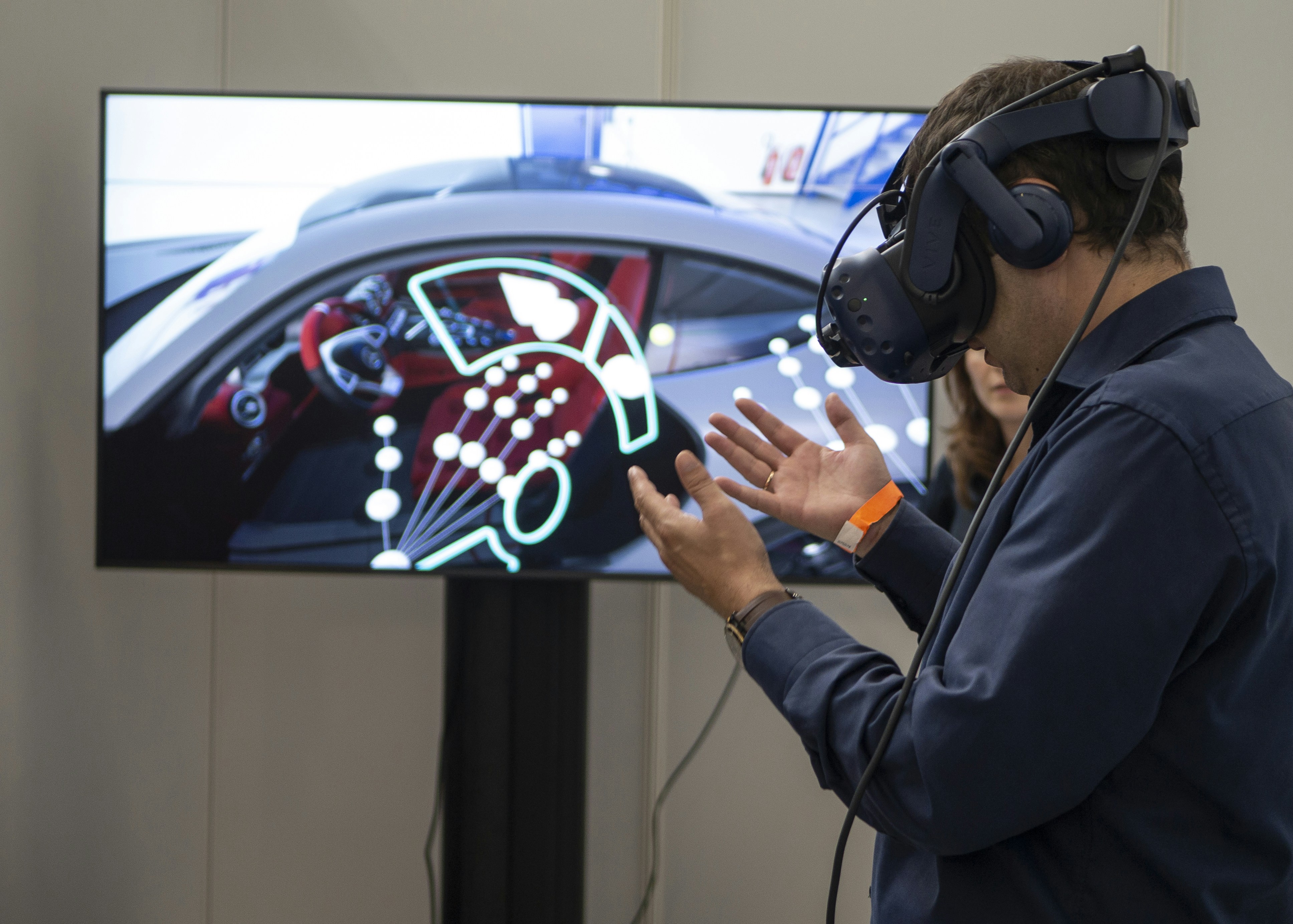 An attendee tries a virtual reality headset, moving their hands in front of a monitor showing a car simulation.