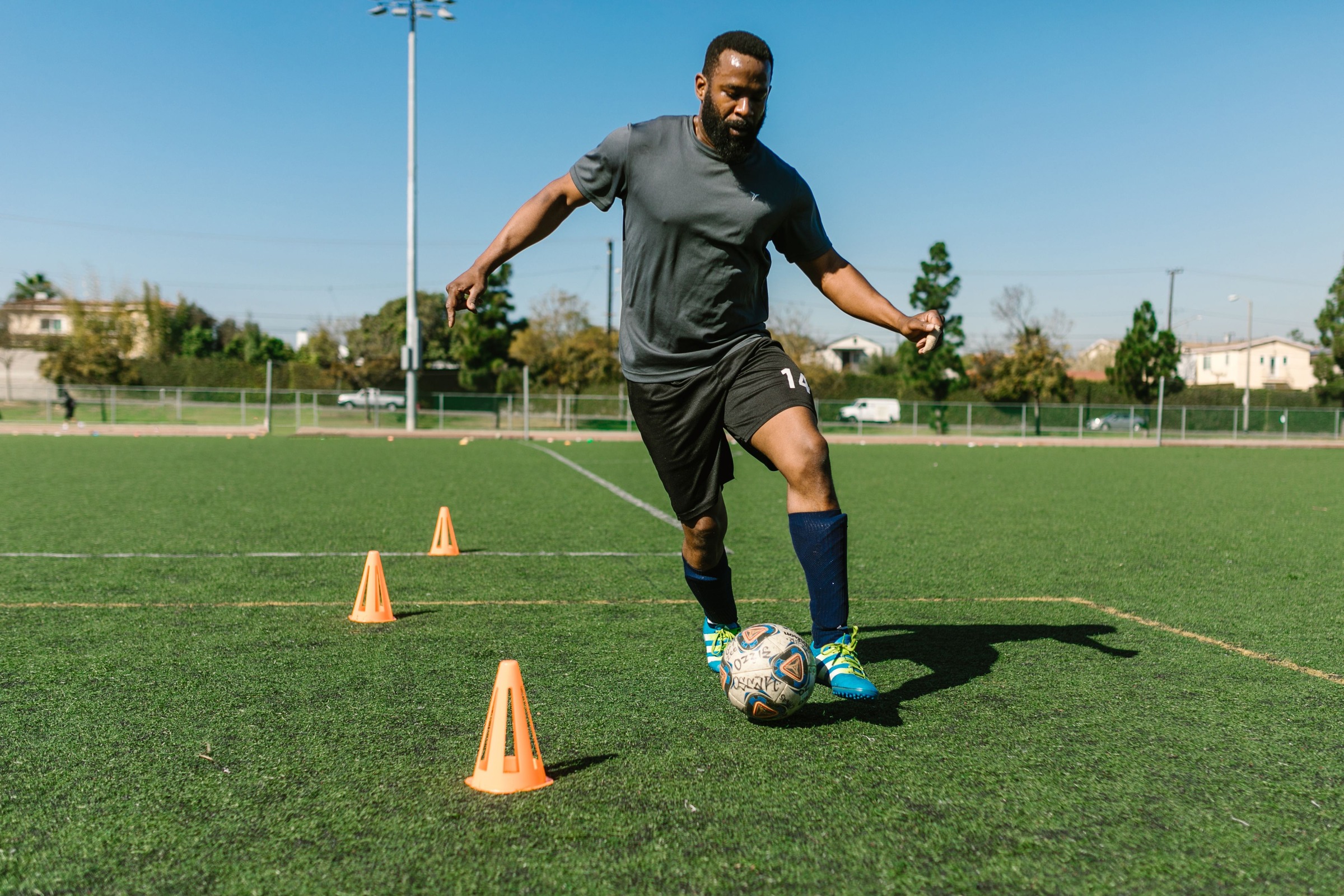 An adult controlling a soccer ball between orange training cones on an outdoor field.