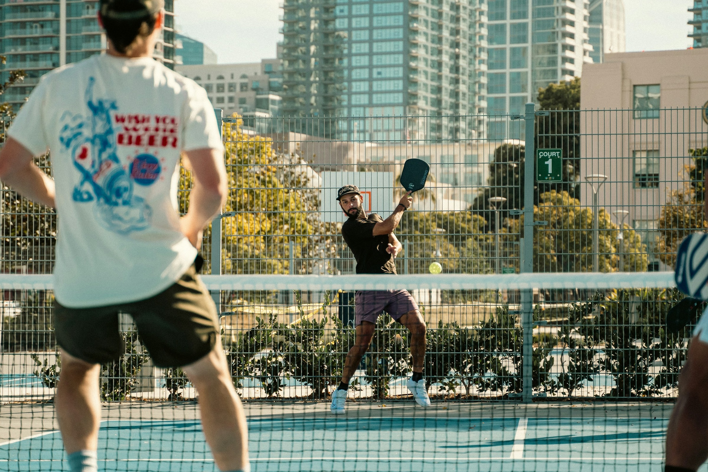 Pickleball players rally across a net on an outdoor court, with city buildings behind them.